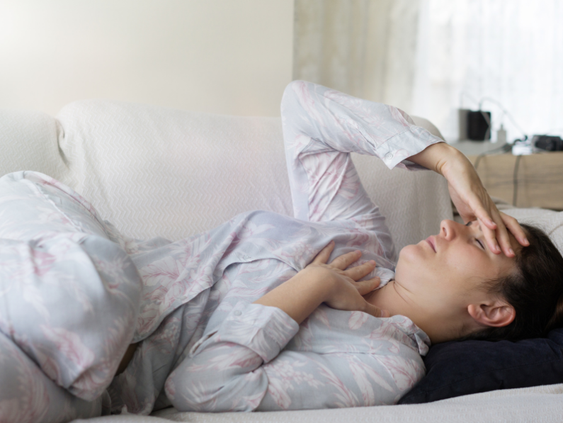 woman lying on couch in pajamas with hand over chest and other hand on head in pain
